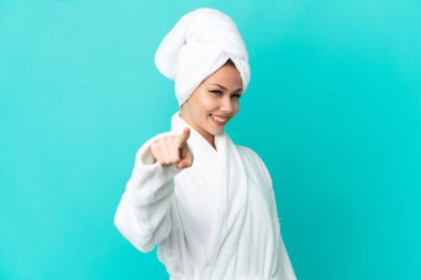 İzole edilmiş mavi arka planda bornozlu genç sarışın kız mutlu bir ifadeyle öne bakıyor.