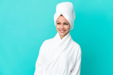 İzole edilmiş mavi arka planda bornozlu genç sarışın kız yana bakıyor ve gülümsüyor.