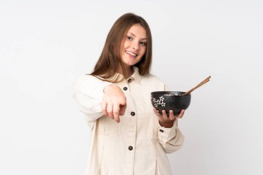 Beyaz arka planda izole edilmiş beyaz tenli genç bir kız elinde çubukla elinde bir kase erişte tutarken kendine güvenen bir ifadeyle seni işaret ediyor.