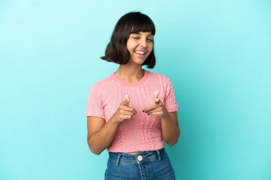 Mavi arka planda izole edilmiş genç melez kadın ön tarafı işaret ediyor ve gülümsüyor.