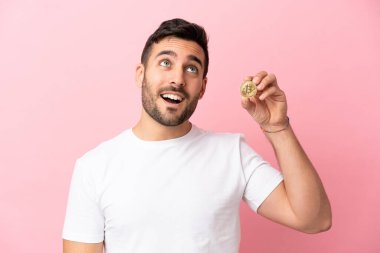Elinde bir Bitcoin tutan genç adam pembe arka planda izole edilmiş bir fikir ararken bir yandan da