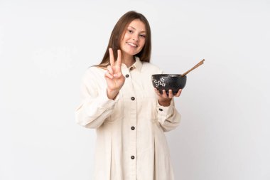 Beyaz arka planda izole edilmiş beyaz bir genç kız elinde çubuklarla elinde bir kase erişte tutarken zafer işareti gösteriyor.