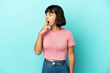 Genç melez kadın mavi arka planda izole edilmiş esniyor ve ağzı açık bir şekilde elleriyle ağzını kapatıyor.