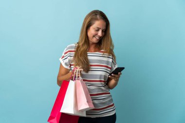 Orta yaşlı Brezilyalı bir kadın mavi arka planda izole edilmiş alışveriş torbaları tutuyor ve bir arkadaşına cep telefonuyla mesaj yazıyor.