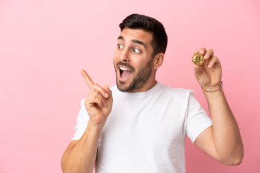 Elinde pembe arka planda izole edilmiş bir Bitcoin tutan genç adam çözümü bulmak için parmağını kaldırıyor.