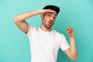 Mavi arka planda izole edilmiş bir Bitcoin tutan genç adam yan tarafa bakarken sürpriz jest yapıyor.