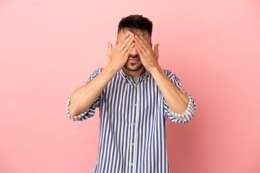 Pembe arka planda izole edilmiş genç, beyaz bir adam gözlerini elleriyle kapatıyor.