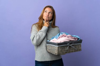 Orta yaşlı bir kadın mor arka planda izole edilmiş çamaşır sepetini tutarken yukarı bakarken şüpheleri oluyor.