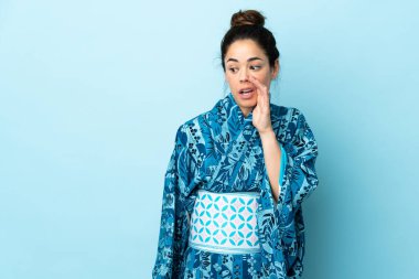 Woman wearing kimono over isolated background whispering something with surprise gesture while looking to the side