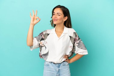 İzole edilmiş mavi arka plan üzerinde genç bir kadın parmaklarıyla tamam işareti gösteriyor.