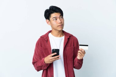 Genç Çinli bir adam beyaz arka planda izole edilmiş. Cep telefonuyla kredi kartıyla alışveriş yapıyor.