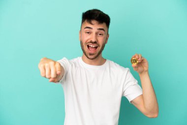 Elinde mavi arka planda izole edilmiş Bitcoin tutan genç adam şaşırmış ve ön tarafı işaret ediyor.