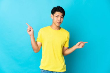 Mavi arka planda izole edilmiş genç Çinli adam parmak uçlarını işaret ediyor ve mutlu