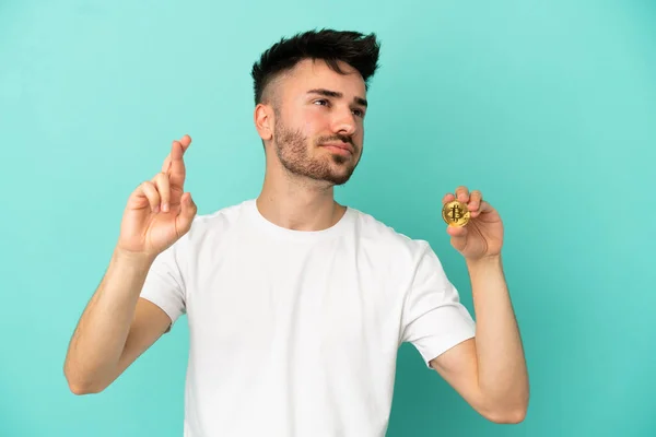 Młody Człowiek Trzyma Bitcoin Odizolowany Niebieskim Tle Palcami Skrzyżowania Życząc — Zdjęcie stockowe