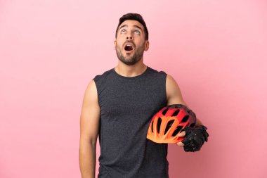 Pembe arka planda izole edilmiş genç bisikletçi yukarı bakıyor ve şaşırmış bir ifadeyle