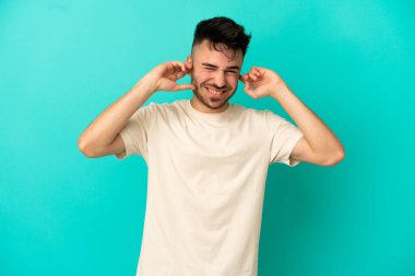 Mavi arka planda izole edilmiş genç beyaz adam hayal kırıklığına uğramış ve kulaklarını kapatmış.