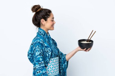 İzole edilmiş beyaz arka planda kimono giyen bir kadın elinde çubuklarla bir kase şehriye tutarken mutlu bir ifade takınıyor.