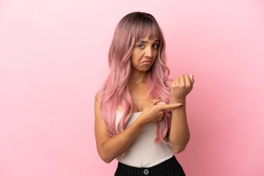 Pembe saçlı, melez bir kadın pembe arka planda, geç kalma jesti yapıyor.