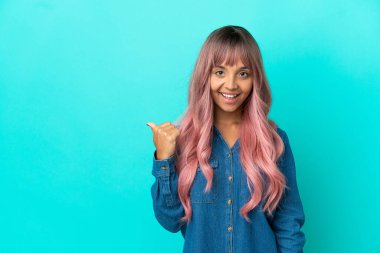 Mavi arka planda izole edilmiş pembe saçlı melez bir kadın bir ürünü sunmak için kenarı işaret ediyor.