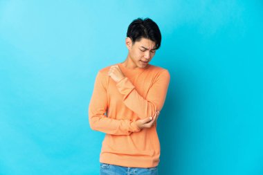 Genç Çinli bir adam mavi arka planda izole edilmiş, dirseğinde ağrı var.