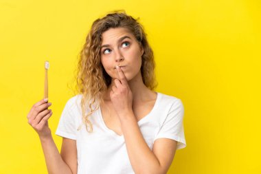 Sarı arka planda dişlerini fırçalayan beyaz bir kadın yukarı bakarken şüpheleri var.