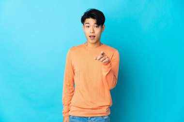 Mavi arka planda izole edilmiş genç bir Çinli şaşırmış ve ön tarafı işaret ediyor.