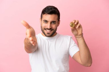 Pembe arka planda izole edilmiş Bitcoin tutan genç adam iyi bir anlaşma için el sıkışıyor.