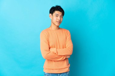 Genç Çinli bir adam mavi arka planda izole edilmiş, kolları bağlı ve ileriye bakıyor.
