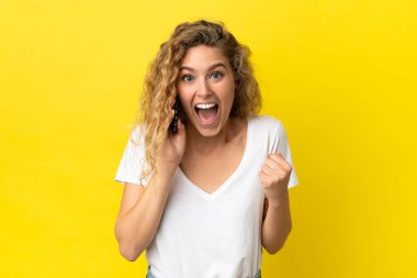 Sarı arka planda cep telefonu kullanan genç sarışın kadın zaferi kutluyor.