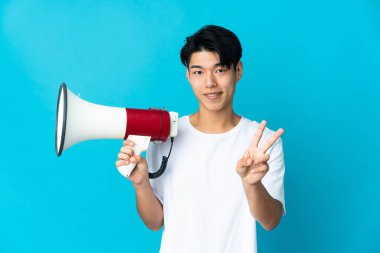 Mavi arka planda izole edilmiş genç bir Çinli elinde bir megafon, gülümsüyor ve zafer işareti gösteriyor.
