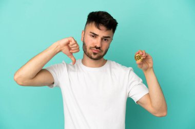 Elinde mavi arka planda izole edilmiş bir Bitcoin tutan genç adam negatif ifadeyle baş parmağını aşağı gösteriyor.