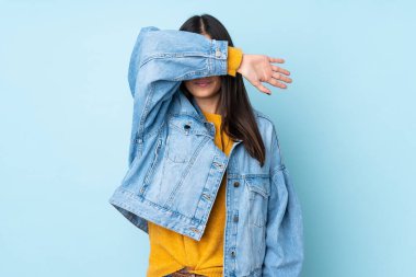 Mavi arka planda izole edilmiş genç beyaz kadın gözlerini elleriyle kapatıyor.