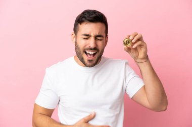 Pembe arka planda izole edilmiş Bitcoin tutan genç adam, çok gülümsüyor.