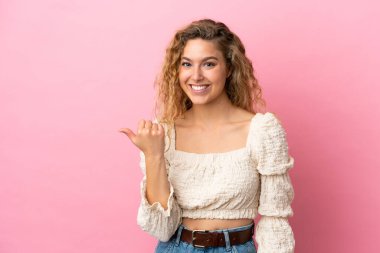 Pembe arka planda izole edilmiş genç sarışın kadın bir ürünü sunmak için kenarı işaret ediyor.