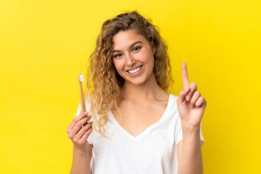 Sarı arka planda dişlerini fırçalayan beyaz bir kadın en iyinin işareti olarak parmağını kaldırıyor.