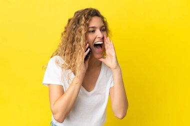 Sarı arka planda izole edilmiş cep telefonu kullanan genç sarışın kadın ağzı sonuna kadar açık bağırıyor.