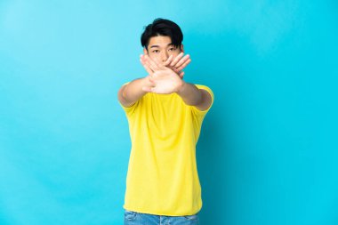 Genç Çinli bir adam mavi arka planda izole edilmiş bir şekilde bir eylemi durdurmak için eliyle jest yapıyor.
