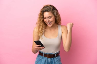 Pembe arka planda izole edilmiş genç sarışın kadın cep telefonu kullanıyor ve zafer jesti yapıyor.
