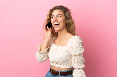 Pembe arka planda izole edilmiş genç sarışın kadın cep telefonuyla sohbet ediyor.