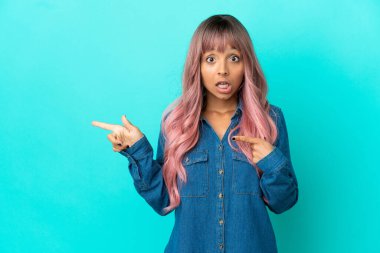 Mavi arka planda izole edilmiş pembe saçlı melez kadın şaşırmış ve işaret ediyor.