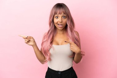 Pembe saçlı melez bir kadın. Pembe arka planda izole edilmiş. Şaşırmış ve işaret ediyor.