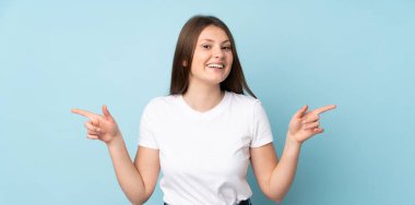 Mavi arka planda izole edilmiş genç beyaz kız parmak uçlarını işaret ediyor ve mutlu.