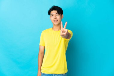 Mavi arka planda izole edilmiş genç Çinli adam gülümsüyor ve zafer işareti gösteriyor.