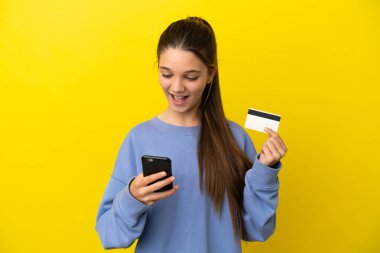 Küçük kız sarı arka planda kredi kartıyla cep telefonuyla alışveriş yapıyor.