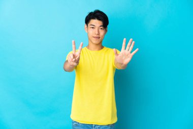 Mavi arka planda izole edilmiş genç Çinli adam yediyi parmaklarıyla sayıyor