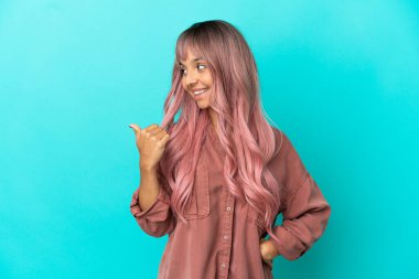 Mavi arka planda izole edilmiş pembe saçlı melez bir kadın bir ürünü sunmak için kenarı işaret ediyor.
