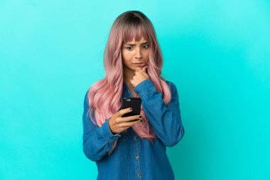 Pembe saçlı genç melez kadın mavi arka planda izole edilmiş düşünüyor ve bir mesaj gönderiyor.