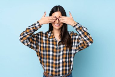 Mavi arka planda izole edilmiş genç beyaz bir kadın. Gözleri elleriyle kaplıyor ve gülümsüyor.