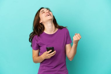 Genç beyaz kadın mavi arka planda izole edilmiş ve telefon zafer pozisyonunda.