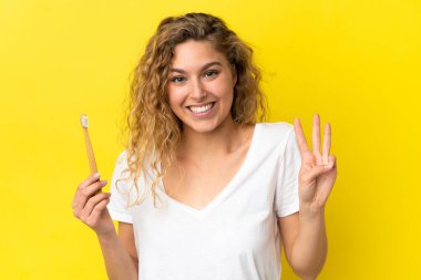 Sarı arka planda dişlerini fırçalayan beyaz bir kadın mutlu ve parmaklarıyla üçe kadar sayıyor.
